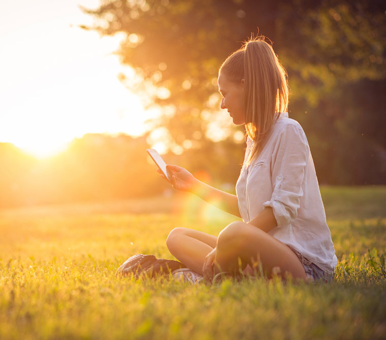 Junge Frau sitzt im Sonnenuntergang auf einer Wiese und betrachtet ihr Mobiltelefon